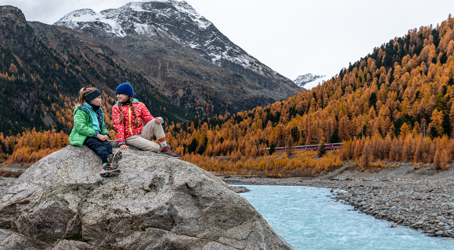 Zwei Kinder in namuk-Kleidung sitzen auf einem Stein inmitten der Engadiner Natur