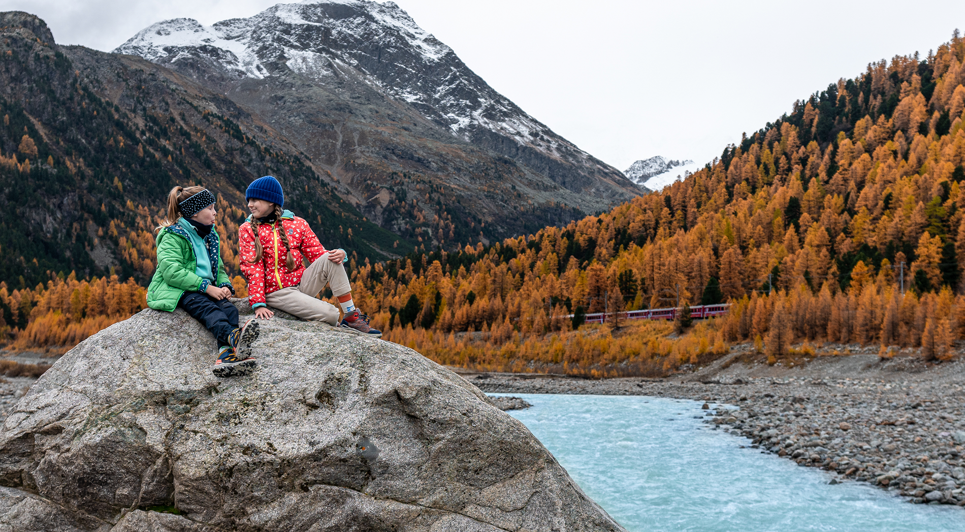 Zwei Kinder in namuk-Kleidung sitzen auf einem Stein inmitten der Engadiner Natur